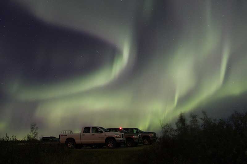 Aurora borealis swirling above parked trucks at night.